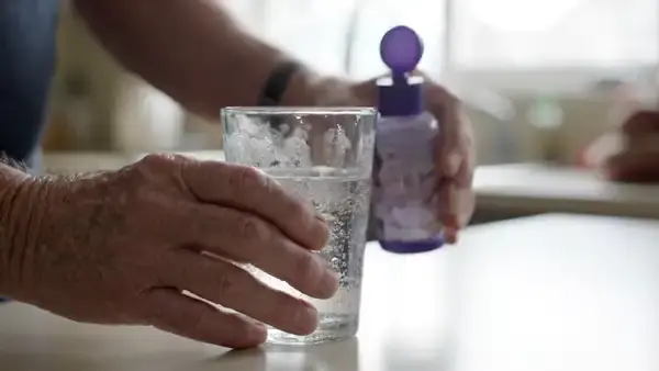 A man's hands squeezing a small purple bottle once over a tall glass of water and swirling it